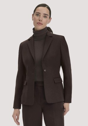 Frau trägt einen dunkelbraunen Blazer und Rollkragenpullover mit passenden Hosen und steht vor einem einfarbigen Hintergrund.