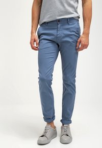 Ljusblå, slim-fit chinos med en jämn textur, som har en brun knappstängning och uppvikta ärmslut, i kombination med ljusgrå sneakers.