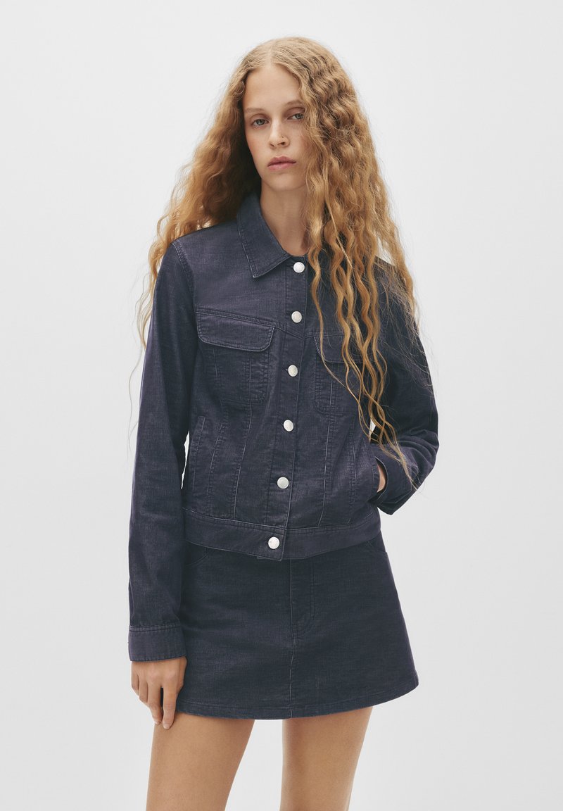Junge Frau mit langen, lockigen Haaren, die eine dunkle Denimjacke und einen passenden Rock trägt, steht vor einem schlichten, hellen Hintergrund.