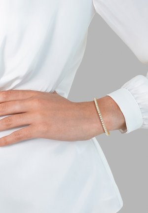 Bracelet en or avec une rangée de pierres claires et scintillantes disposées sur la surface. Porte sur un poignet contre une blouse blanche. Design simple et élégant.