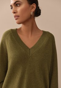 Green v-neck sweater in soft knit material with ribbed accents. Model wears large hoop earrings, showcasing a close-up view of the garment.