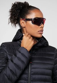 Una persona con un piumino nero, che indossa occhiali da sole dal design elegante a forma di avvolgimento, con accenti rossi e lenti scure, che mostrano uno stile sportivo.