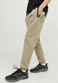 Beige sweatpants with a tapered fit, featuring stitched detailing and a side zip pocket, paired with black athletic shoes and white socks.