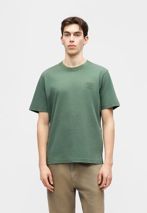 Junger Mann trägt grünes T-Shirt mit kurzen Ärmeln und kleinem schwarzem Logo auf der Brust sowie hellbraune Hose, steht vor einfarbig hellem Hintergrund.