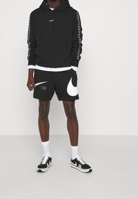 Czarna bluza Nike z białym logo na klatce piersiowej, czarne szorty z dużym białym swooshem i szarym logo TM, w połączeniu z czarno-białymi sneakersami.