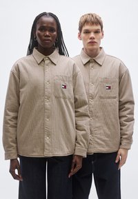 Beige kockás gombos ingek laza szabással, folt zsebekkel és bal oldali mellkasi logóval. Két modell viseli, akik egymás mellett állnak.