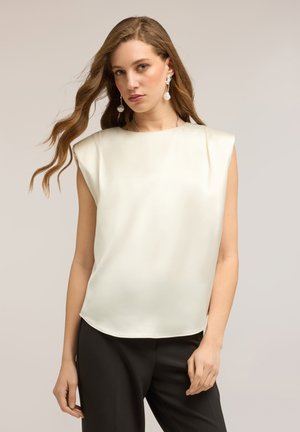 Giovane donna con capelli lunghi e ondulati, che indossa un top in satin bianco senza maniche e pantaloni neri su uno sfondo semplice.