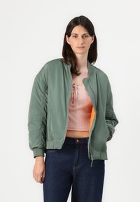 Chaqueta bomber verde con puños acanalados, cierre de cremallera y forro naranja, llevada sobre una camiseta acanalada rosa y jeans oscuros.