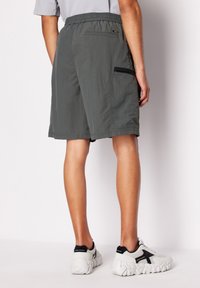 Pantaloni corti grigi con una cintura elastica, una tasca con zip sul lato destro e un tessuto liscio e leggero. Indossati con scarpe da ginnastica bianche.