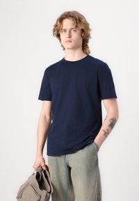Camiseta de cuello redondo azul marino hecha de algodón, combinada con jeans de pernera ancha de lavado claro. El modelo sostiene una bolsa tote gris, mostrando un diseño simple.
