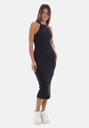 Femme portant une robe midi noire sans manches et des baskets compensées blanches, debout devant un fond clair uni.