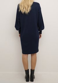 Abito blu navy lungo fino al ginocchio con maniche lunghe a sbuffo, polsini a coste e silhouette aderente. Tessuto liscio, indossato con stivaletti neri con tacco.