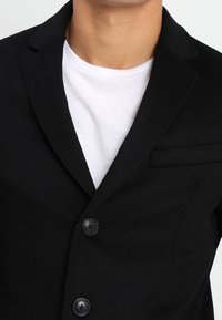 Une personne portant un blazer noir par-dessus un t-shirt blanc, mettant l'accent sur le haut du torse et les boutons du blazer. Fond neutre.