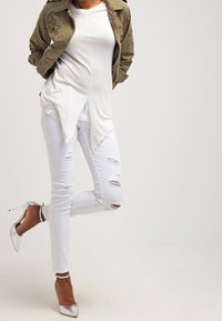 Camiseta blanca de corte largo con dobladillo en punta, jeans blancos desgastados, chaqueta verde olivo y zapatos de tacón alto plateados metálicos.