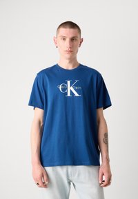 Camiseta de algodón azul con mangas cortas y cuello redondo. Presenta un gran logo blanco "CK" en el centro. Tejido texturizado, ajuste casual.