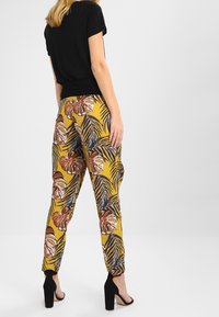 Un haut noir à manches courtes associé à un pantalon jaune à motifs floraux, mettant en avant de grandes feuilles et fleurs. Des chaussures à talons noirs complètent le look.