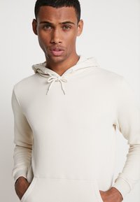 Lys beige hettegenser med kengurulomme, snorhette og ribbestrikkede mansjetter. Laget av myk, teksturert stoff. Minimalistisk design.