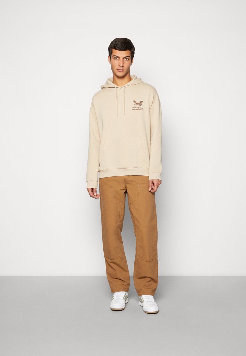 Felpa beige con cappuccio, dotata di tasca frontale e design stampato. Abbinata a pantaloni larghi color sabbia e sneakers bianche con dettagli colorati.