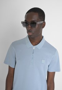 Lichtblauw polo shirt met een rits en een klein logo op de borst. Model draagt een bril met een doorzichtige montuur, met vierkante lenzen.