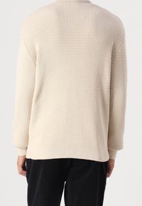 Pull en tricot beige avec un motif texturé, manches longues et ourlet côtelé ; doté d'un col rond et d'une coupe décontractée.