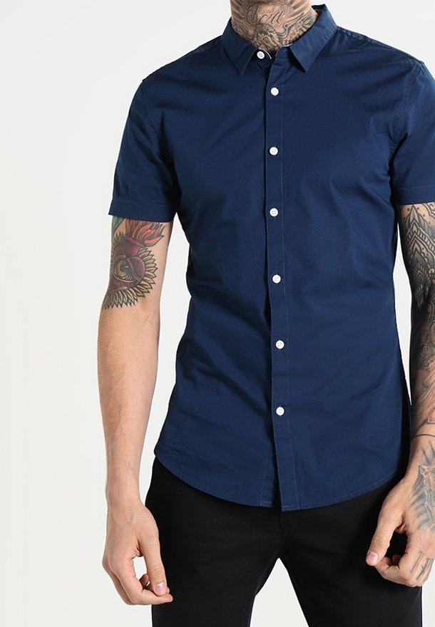 Uomo che indossa una camicia a maniche corte blu navy aderente e pantaloni neri, con tatuaggi su entrambe le braccia e sul collo.