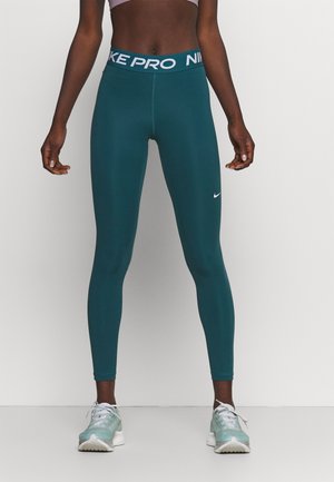 Femme portant des leggings Nike Pro teal et des chaussures de course blanches, debout contre un fond blanc uni.