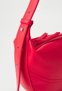 Red faux leather handbag with a curved shape, a long adjustable strap, and a zipper closure. Features soft texture and minimalistic design details.