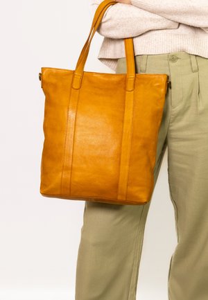 Keltainen nahkainen olkalaukku, jossa on kaksi tukevata kahvaa, sileä pinta ja pystysuorat saumat edessä, nojaa khaki-housuihin.