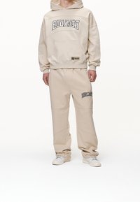 Felpa in cotone beige con tasca frontale e testo grafico nero, abbinata a pantaloni della tuta beige coordinati con dettagli laterali e logo.