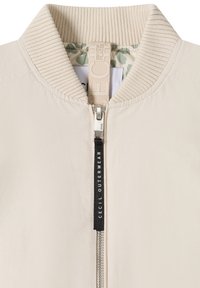 Blouson aviateur beige avec col côtelé, fermeture éclair intégrale à l'avant et doublure à motif floral visible à l'intérieur du col.