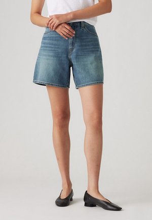 Frau trägt blaue Jeansshorts, weißes Top und schwarze Schuhe mit niedrigem Absatz, steht mit verschränkten Händen vor einem einfarbigen Hintergrund.
