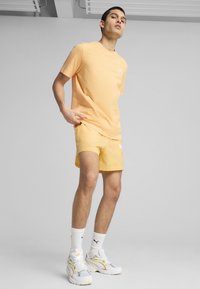 T-shirt a maniche corte color pesca abbinato a pantaloni corti dello stesso colore, entrambi realizzati in tessuto leggero. Footwear comprende sneaker bianche con dettagli gialli.
