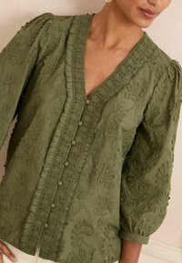 Blouse vert olive avec broderie florale texturée, décolleté en V, fermeture à boutons et manches bouffantes. Fabriquée à partir d'un tissu doux et léger.