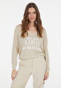 Beige Langarm-V-Ausschnitt-Oberteil mit weißem Text: "REBELLE MIT EINEM LÄCHELN, SCHWEBEND MIT GESCHLOSSENEN AUGEN." Weicher, strukturierter Stoff.