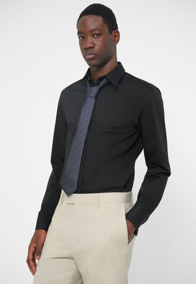 Μαύρη μακρυμάνικη πουκαμίσα με γωνιαστό γιακά, συνδυασμένη με φωτεινά μπεζ ξεχωριστά παντελόνια και ναυτικό γραβάτα με διαγώνιες ρίγες.