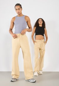 Två modeller bär avslappnade kläder som inkluderar beige, snörade sweatpants och figurnära crop tops i svart och ljusgrått. Släta, mjuka texturer.