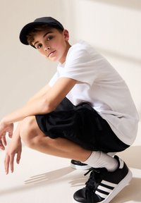 Juoda kepurė, balti marškinėliai, juodos trumpikės ir juodi sportbačiai su baltais akcentais. Lygios tekstūros ir kasdieniškas sportiškas dizainas.