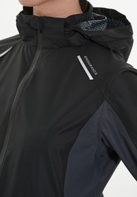 Svart vattentät jacka med hög krage, reflekterande detaljer och blåmönstrad insida. Har en hel dragkedja och sportig design.