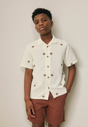 Teenager-Junge trägt ein weißes Kurzarmhemd mit braunen Rautenmuster und braune Shorts, steht vor einer schlichten beigen Wand.