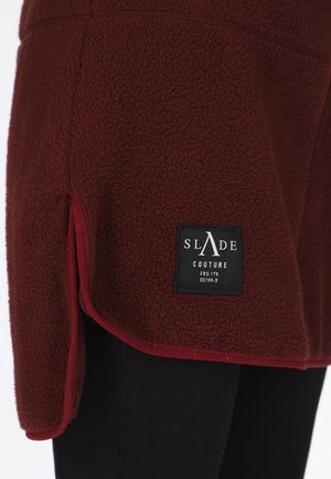 Rode fleece top met een gestructureerde afwerking, voorzien van een zwart label met witte tekst. Randen omrand met een contrasterende kleur. Korte lengte.