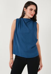Modrý top bez rukávů s vysokým výstřihem, splývavým materiálem, texturovaným povrchem a volným střihem, doplněný o černé kalhoty a jednoduché stříbrné šperky.