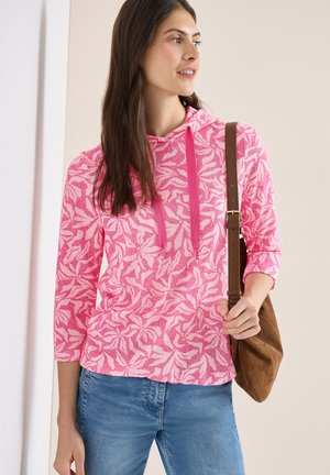 Femme portant un sweat à capuche rose à motifs avec des manches trois-quarts, un jean bleu et un sac à bandoulière marron, regardant sur le côté en intérieur.