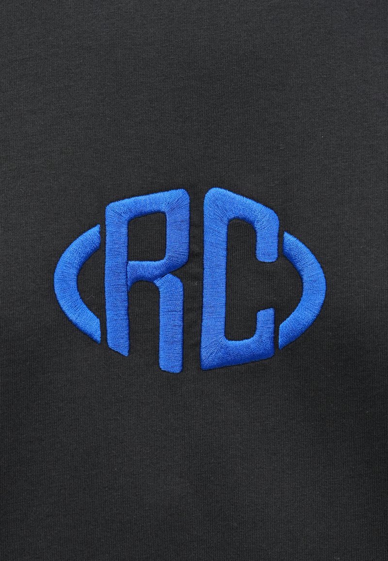 Blaue gestickte Buchstaben "RC" mit geschwungenen Linien auf beiden Seiten auf einem schwarzen Stoffhintergrund.