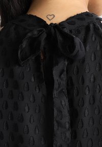 Mustasilkkinen yläosa, jossa on kohokuvioitu pisaramainen kuvio. Takana on rusetti kiinnitys ja kaulassa pieni sydämetatuointi.