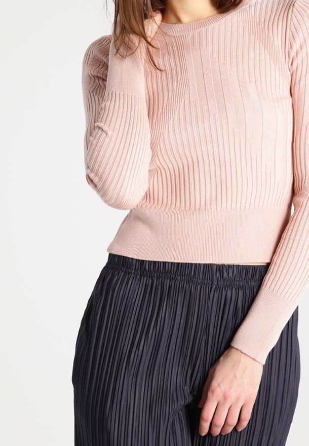 Femme portant un pull en maille côtelée rose clair à manches longues et un pantalon plissé foncé taille haute, une main touchant ses cheveux.