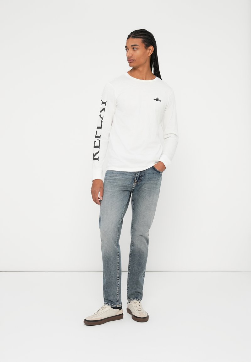 T-shirt bianco a maniche lunghe con la scritta "REPLAY" stampata in nero sulla manica sinistra, abbinata a jeans azzurri chiari e sneaker color crema.