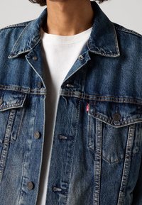 Jeansjacka i mörkblått med lätt slitage, knäppning framtill, två bröstfickor och en liten röd logotypetikett på övre vänstra sidan.