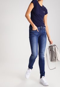 Nainen pukeutunut tummansiniseen t-paitaan, istuviin sinisiin farkkuihin, valkoisiin lenkkareihin, pitäen oikeassa kädessään olkahihnalla varustettua vaaleanharmaata käsilaukkua.