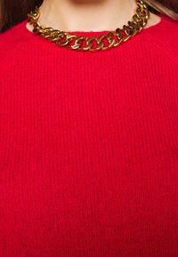 Punainen neulottu paita, jossa on teksturoitu pinta, yhdistetty paksuun kultaketjuun. Kaula-aukko on pyöreä ja siinä on kevyttä ribbineulosta.