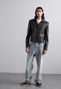 Black leather biker jacket with silver zippers, worn over a white shirt; paired with light blue baggy jeans and black shoes.